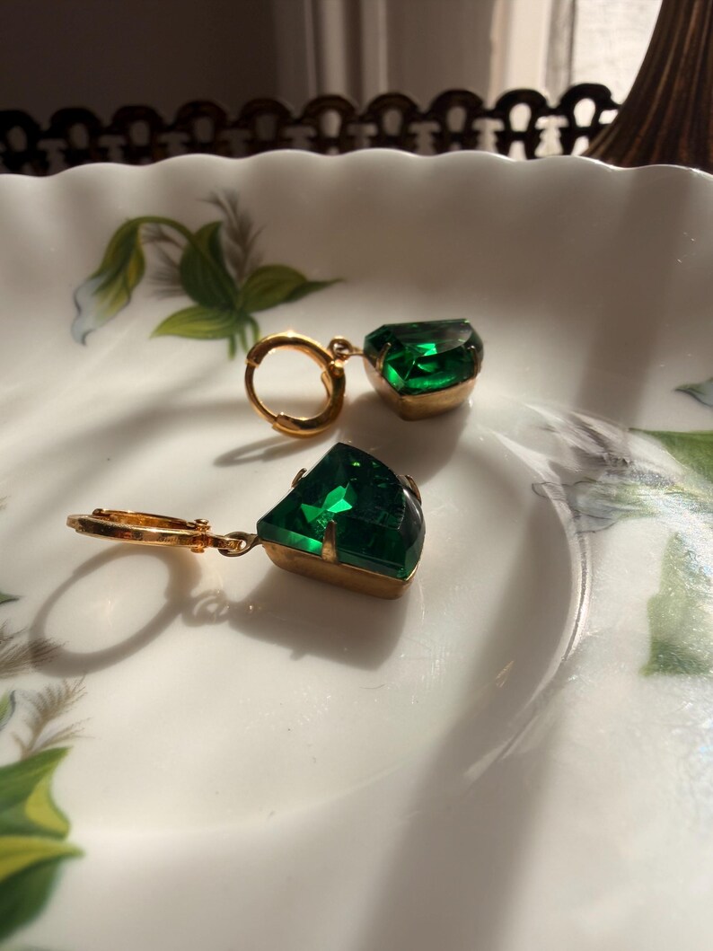 May include: A pair of gold-tone hoop earrings featuring faceted, emerald-green, trapezoidal-shaped stones set in metal prongs. The earrings are designed with a hinged closure mechanism, showcasing a vibrant green hue and a polished metallic finish.