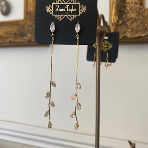 May include: Gold dangle earrings with a delicate branch design featuring clear marquise-shaped crystals. The earrings are displayed on a black card with the text "Zara Taylor" printed on it.