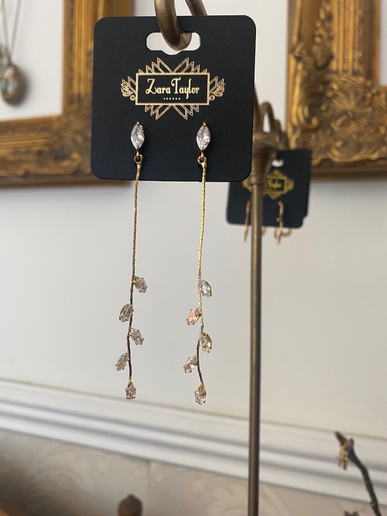 May include: Gold dangle earrings with a delicate branch design featuring clear marquise-shaped crystals. The earrings are displayed on a black card with the text "Zara Taylor" printed on it.