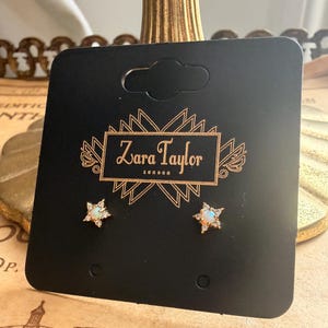 May include: A pair of gold star-shaped stud earrings featuring a central iridescent opal stone surrounded by small clear crystals. The earrings are mounted on a black display card with the gold text Zara Taylor London printed in the center.