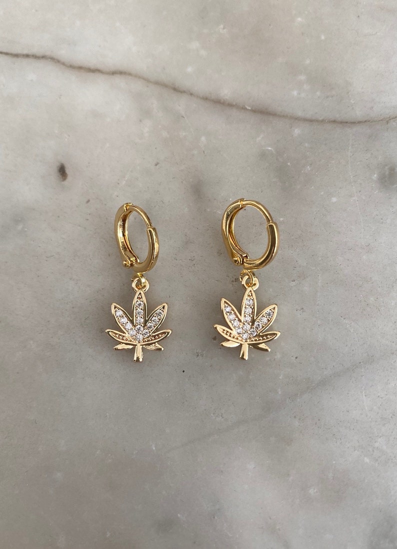 May include: A pair of gold-colored hoop earrings. Each earring features a small, gold-colored cannabis leaf pendant, embellished with clear, sparkling crystals. The earrings are displayed against a light gray background.
