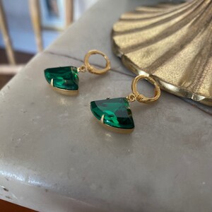 May include: A pair of gold hoop earrings featuring faceted, fan-shaped emerald green stones. Each earring consists of a small circular gold hoop attached to a triangular, curved green gemstone set in a gold metal frame, creating a dangle design.