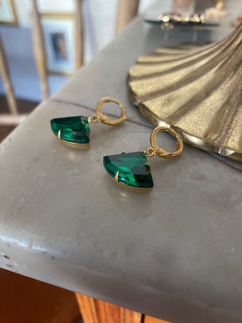 May include: A pair of gold hoop earrings featuring faceted, fan-shaped emerald green stones. Each earring consists of a small circular gold hoop attached to a triangular, curved green gemstone set in a gold metal frame, creating a dangle design.