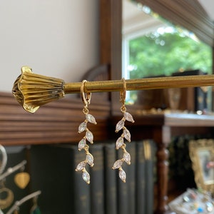 May include: A pair of gold-colored leaf-shaped earrings with clear, sparkling accents. The earrings are hanging from a gold-colored rod. The background includes a mirror and a glimpse of a room.