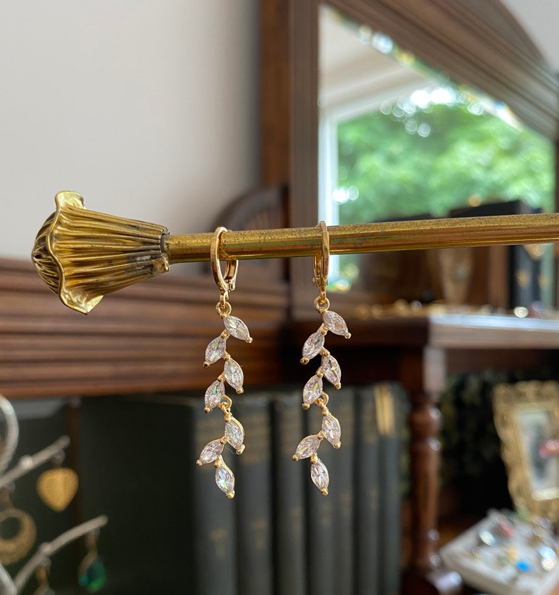 May include: A pair of gold-colored leaf-shaped earrings with clear, sparkling accents. The earrings are hanging from a gold-colored rod. The background includes a mirror and a glimpse of a room.