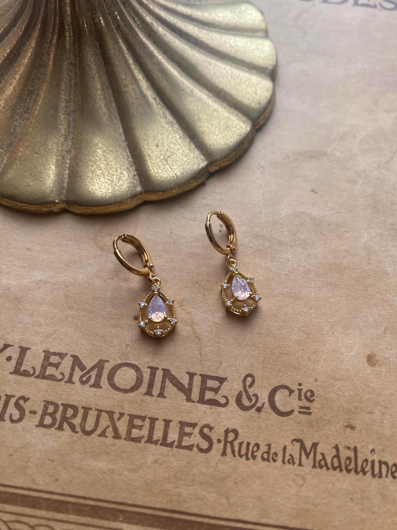 May include: A pair of gold hoop earrings with a teardrop-shaped pendant. Each pendant features a clear crystal with a light pink hue and a gold halo of smaller crystals.