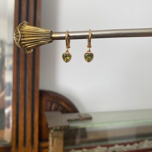 K&ouml;nnte beinhalten: Ein Paar goldene Creolen mit kleinen, herzf&ouml;rmigen gr&uuml;nen Edelsteinen. Jeder Ohrring hat einen zarten, perlenartigen Rand um den herzf&ouml;rmigen Stein, der an einem d&uuml;nnen goldenen Reifen mit einem sicheren Klappverschluss befestigt ist.