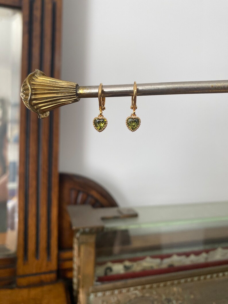 K&ouml;nnte beinhalten: Ein Paar goldene Creolen mit kleinen, herzf&ouml;rmigen gr&uuml;nen Edelsteinen. Jeder Ohrring hat einen zarten, perlenartigen Rand um den herzf&ouml;rmigen Stein, der an einem d&uuml;nnen goldenen Reifen mit einem sicheren Klappverschluss befestigt ist.