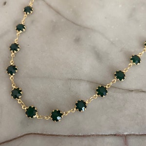 Op de afbeelding: Een gouden ketting met donkergroene ronde stenen. De ketting ligt op een wit marmeren oppervlak.