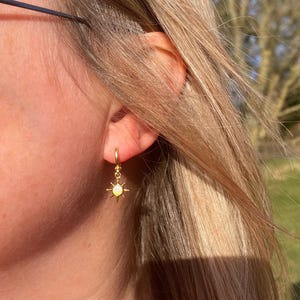 May include: Gold hoop earrings with a small sun charm featuring a yellow opal stone.