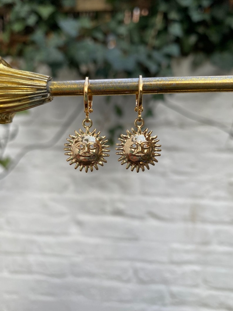 Op de afbeelding: Een paar gouden oorbellen met een zonontwerp met een gezicht. De oorbellen hangen aan een gouden staaf.