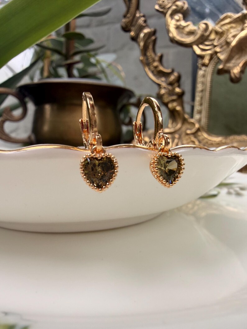 K&ouml;nnte beinhalten: Ein Paar goldfarbene Creolen mit herzf&ouml;rmigen, olivgr&uuml;nen facettierten Steinen. Jeder Stein ist in eine zarte, perlenartige goldfarbene Metallfassung eingefasst und h&auml;ngt an einem kleinen, passenden Klappverschluss in Ringform.