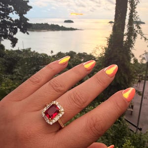 May include: A gold ring featuring a large rectangular red gemstone at the center, surrounded by a halo of small clear stones. The band is also set with small clear stones, and the ring is worn on a finger with bright orange iridescent nails.