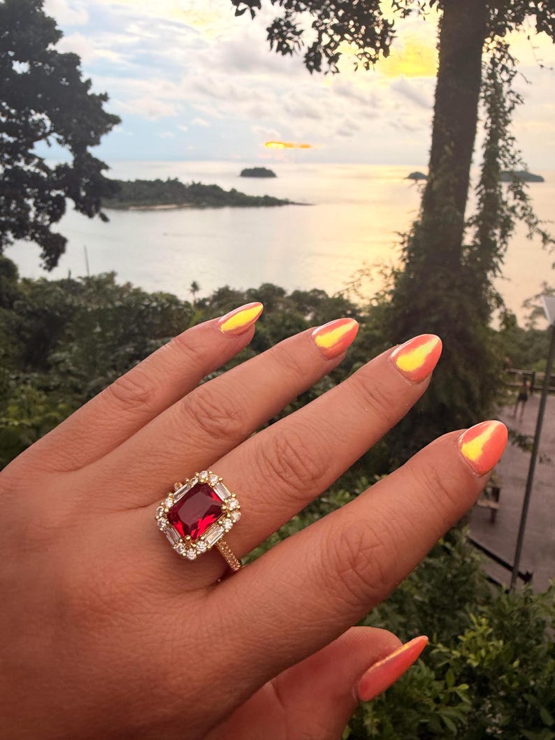 May include: A gold ring featuring a large rectangular red gemstone at the center, surrounded by a halo of small clear stones. The band is also set with small clear stones, and the ring is worn on a finger with bright orange iridescent nails.