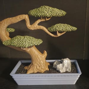 May include: A miniature bonsai tree with a brown, gnarled trunk and green foliage in a light blue rectangular planter. The planter has a decorative border and contains a small gray rock.