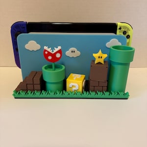 May include: A 3D-printed Nintendo Switch dock featuring a Super Mario Bros. theme. The dock includes a blue backdrop with white clouds, a red and white Piranha Plant, a yellow question mark block, and a green warp pipe. The base has green grass and brown brick details.