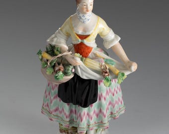 Op de afbeelding: Een porseleinen beeldje van een vrouw in traditionele klederdracht, met een mand met groenten. Het beeldje heeft een gele top, een rood vest, een zwarte rok en een rok met patronen met roze en groene zigzags. De basis is versierd met gouden randen.