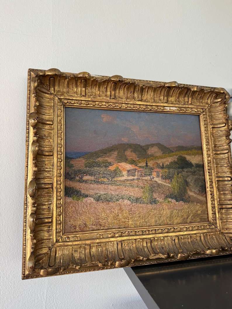 Puede incluir: Una pintura al &oacute;leo enmarcada que representa una escena de paisaje con colinas onduladas, una peque&ntilde;a casa y &aacute;rboles en primer plano. El cielo es de un azul suave con toques de rosa y naranja. La pintura est&aacute; en un marco dorado con detalles ornamentados.