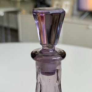 May include: A close-up of a vintage lavender glass bottle with a faceted stopper. The bottle has a slender, fluted design, and the stopper is a darker shade of purple. The glass reflects light, creating a shimmering effect. The bottle is likely for perfume or other liquids.