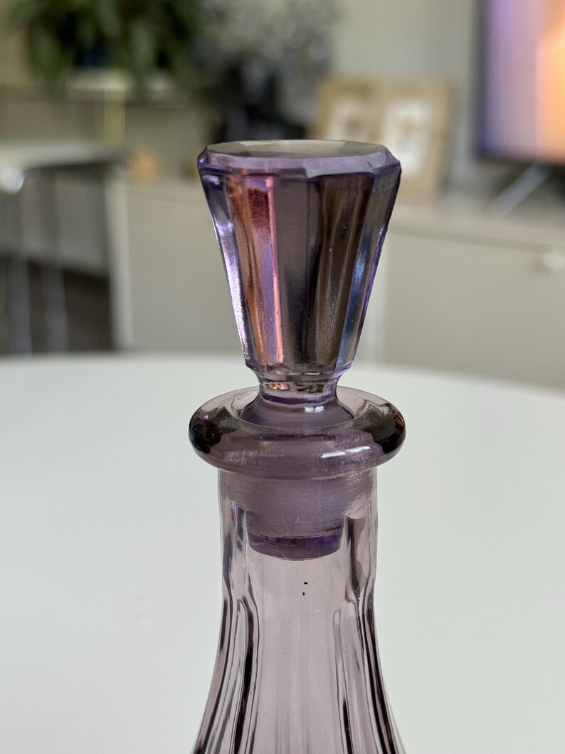 May include: A close-up of a vintage lavender glass bottle with a faceted stopper. The bottle has a slender, fluted design, and the stopper is a darker shade of purple. The glass reflects light, creating a shimmering effect. The bottle is likely for perfume or other liquids.
