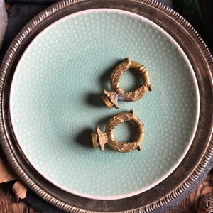 Op de afbeelding: Twee gouden metalen decoratieve eindstukken met sierlijke ontwerpen, liggend op een lichtgroen keramisch bord met een zilveren rand.