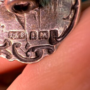 May include: Close-up of a silver button with intricate floral designs. The back of the button has the markings "L&S" and a crown.