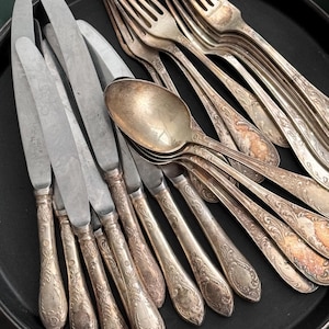 Puede incluir: Un surtido de cubiertos antiguos, que incluye cuchillos, tenedores y cucharas, dispuestos sobre una bandeja negra. La cubertería tiene mangos ornamentados y grabados y hojas y púas de plata deslustrada, lo que sugiere edad y uso. Primer plano.