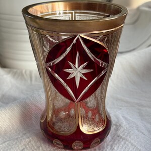 Può includere: Un vaso di vetro rosso con un bordo dorato e intricati disegni di vetro tagliato. Il vaso presenta un design a forma di stella al centro e un motivo a spirale attorno al bordo.