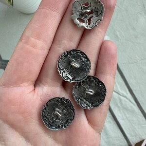 May include: Four silver buttons with intricate designs. The buttons have a raised, ornate border and a small loop on the back for sewing.
