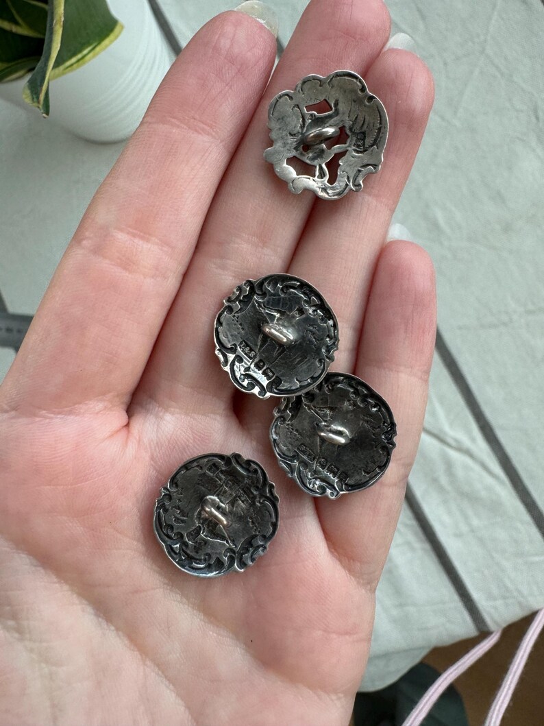 May include: Four silver buttons with intricate designs. The buttons have a raised, ornate border and a small loop on the back for sewing.