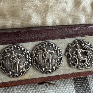 May include: Three silver buttons in a velvet lined box. The buttons feature detailed engravings of figures playing musical instruments.