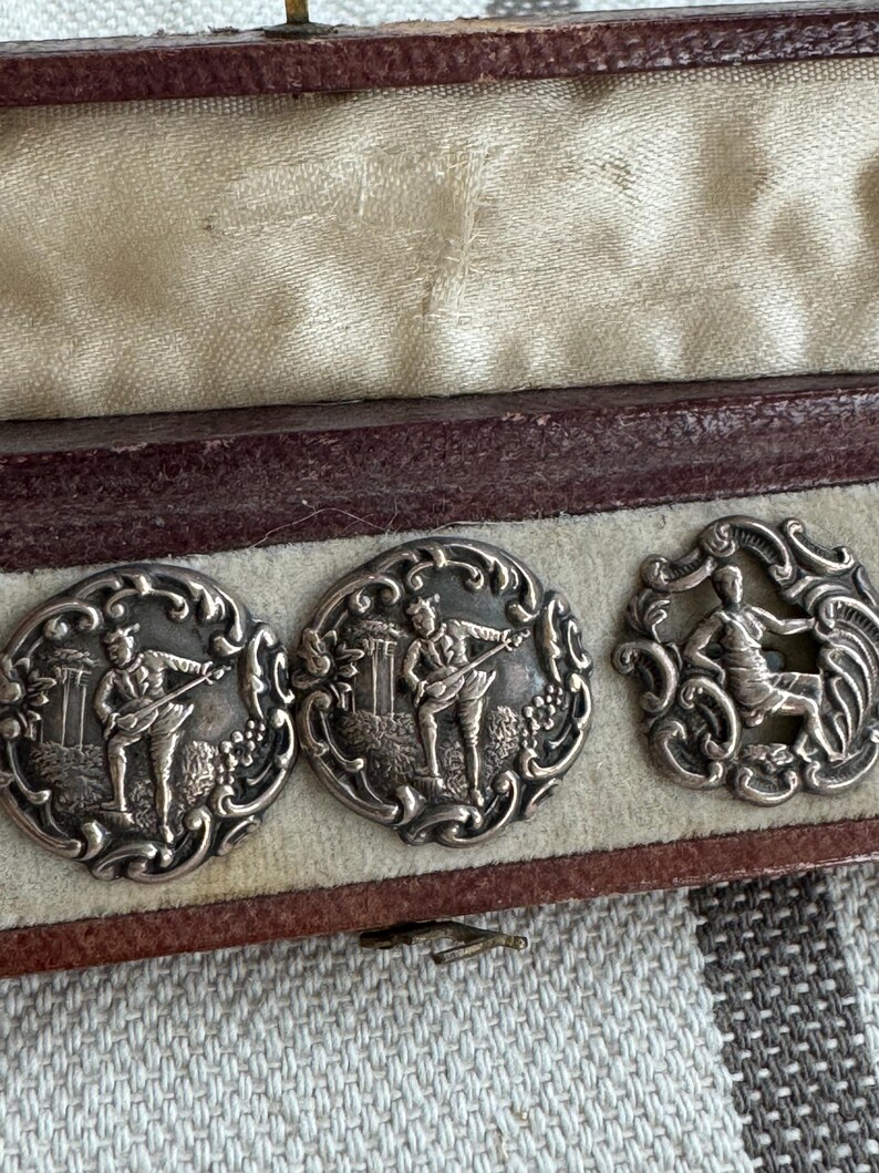 May include: Three silver buttons in a velvet lined box. The buttons feature detailed engravings of figures playing musical instruments.