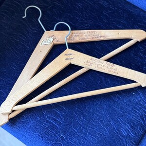 Puede incluir: Dos perchas de ropa de madera vintage con ganchos de metal. Las perchas tienen un acabado de madera natural y presentan el texto "MARCHAND FRERES" y otro texto. Las perchas están sobre una superficie azul.