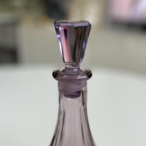 May include: A close-up of a lavender-colored glass bottle with a faceted stopper. The bottle has a slender, elongated shape, and the stopper is a geometric design. The glass has a slight iridescence.