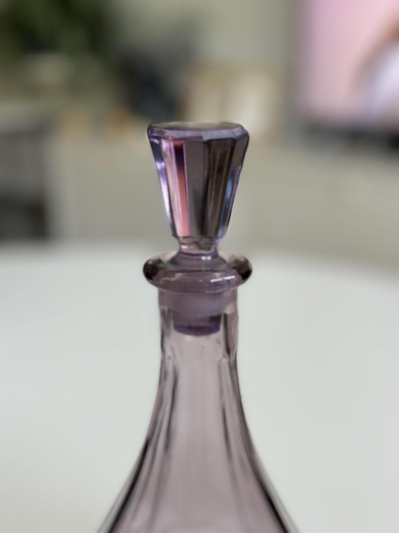 May include: A close-up of a lavender-colored glass bottle with a faceted stopper. The bottle has a slender, elongated shape, and the stopper is a geometric design. The glass has a slight iridescence.