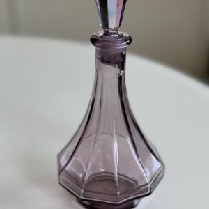 May include: A vintage purple glass perfume bottle with a faceted, geometric design. The bottle has a matching stopper with iridescent accents. The bottle's shape is a slender, elongated cone, and it sits on a hexagonal base.