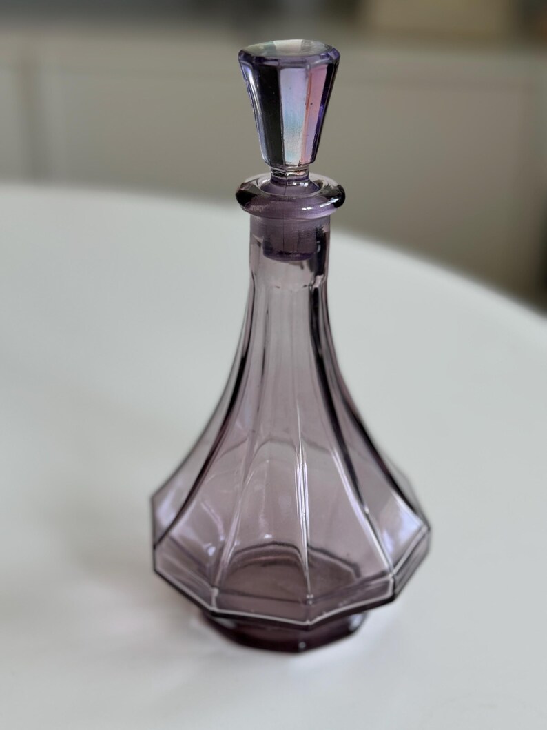 May include: A vintage purple glass perfume bottle with a faceted, geometric design. The bottle has a matching stopper with iridescent accents. The bottle's shape is a slender, elongated cone, and it sits on a hexagonal base.