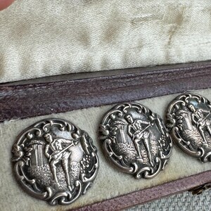 May include: Four silver-toned cufflinks in a brown velvet box. Each cufflink features an intricate design of a man playing a musical instrument in a landscape setting.
