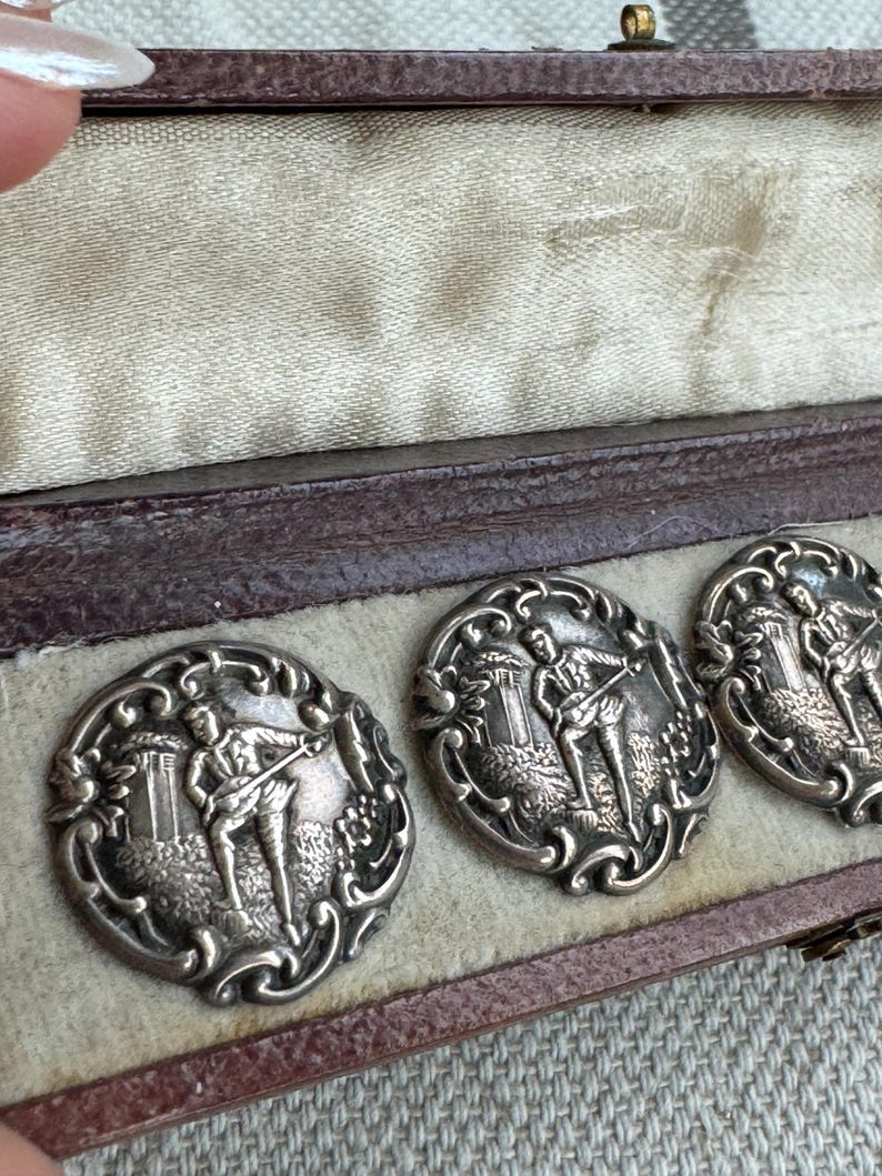 May include: Four silver-toned cufflinks in a brown velvet box. Each cufflink features an intricate design of a man playing a musical instrument in a landscape setting.