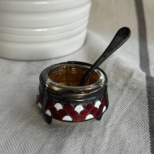 May include: A small, red and white salt cellar with a black rim and three legs. A small silver spoon rests on the rim of the cellar.