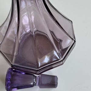 May include: A purple glass perfume bottle with a matching stopper. The bottle has a geometric design with a wide, faceted base and a slender neck. The stopper is rectangular. The glass has a translucent quality.