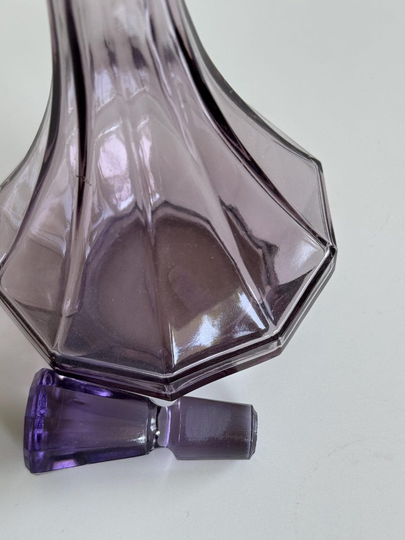 May include: A purple glass perfume bottle with a matching stopper. The bottle has a geometric design with a wide, faceted base and a slender neck. The stopper is rectangular. The glass has a translucent quality.