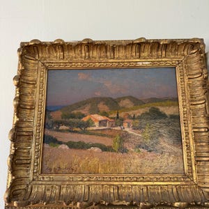 Puede incluir: Una pintura al &oacute;leo enmarcada que representa una escena de paisaje con un peque&ntilde;o pueblo enclavado en colinas onduladas. La pintura est&aacute; ambientada en una luz c&aacute;lida y dorada, con un cielo azul y nubes esponjosas en el fondo. La firma del artista, "Cay de Dainville", es visible en la esquina inferior derecha.