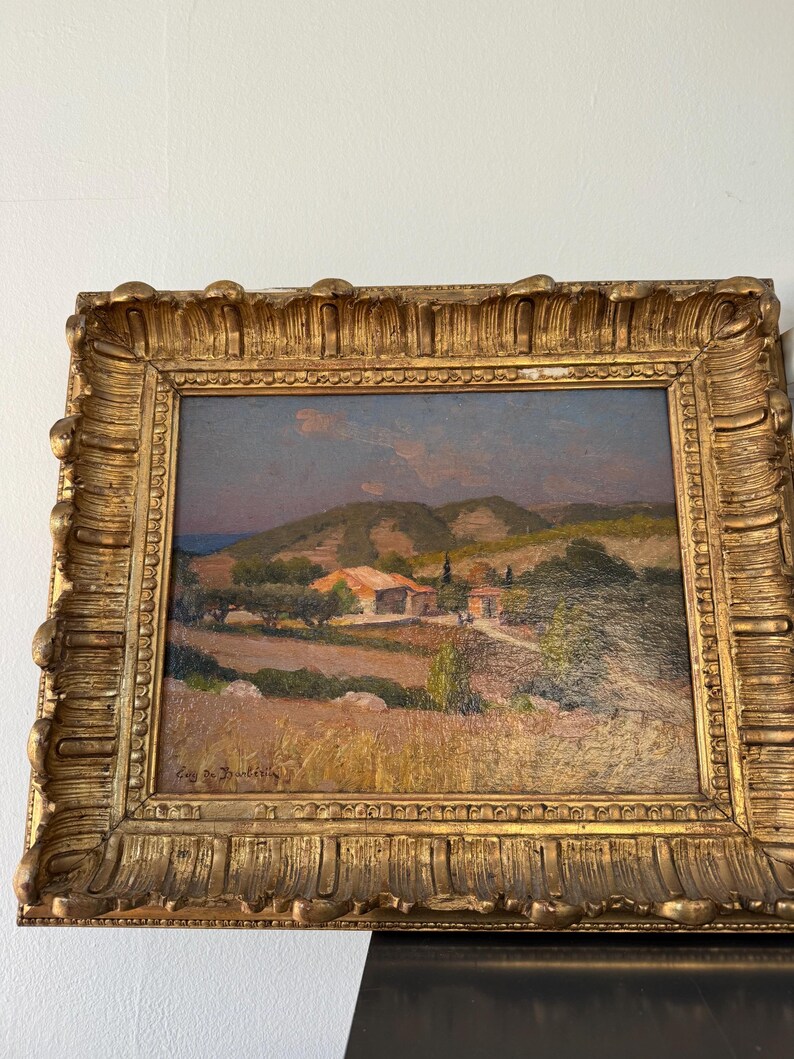 Puede incluir: Una pintura al &oacute;leo enmarcada que representa una escena de paisaje con un peque&ntilde;o pueblo enclavado en colinas onduladas. La pintura est&aacute; ambientada en una luz c&aacute;lida y dorada, con un cielo azul y nubes esponjosas en el fondo. La firma del artista, "Cay de Dainville", es visible en la esquina inferior derecha.