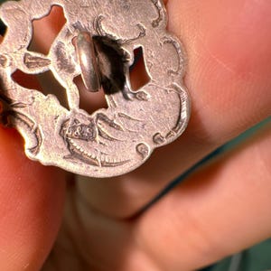 May include: A silver-toned button with an intricate design and a loop on the back. The button has a floral pattern and a raised edge.
