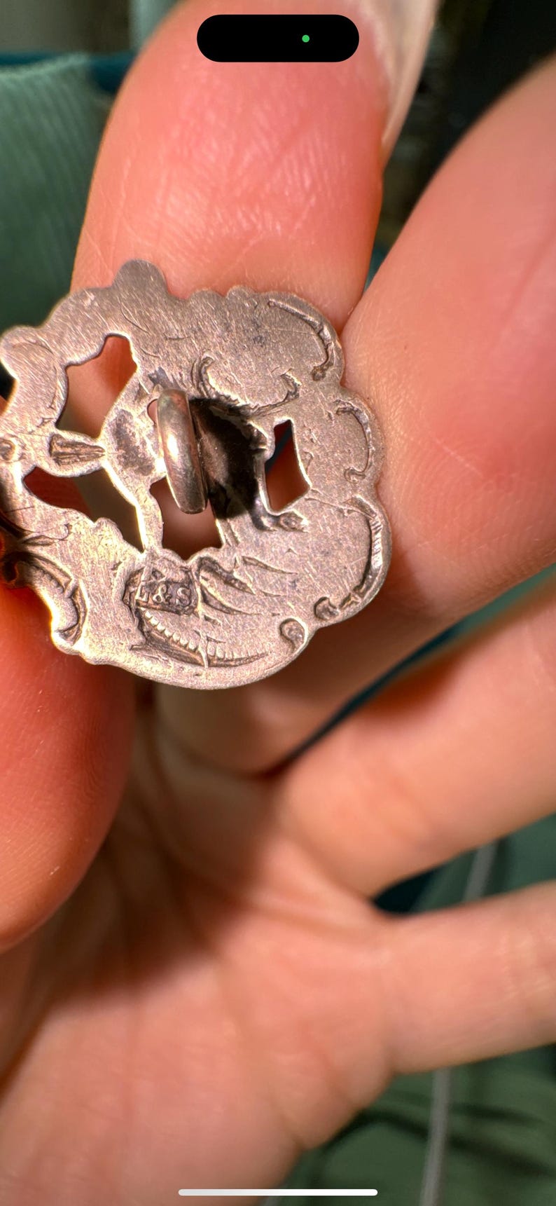 May include: A silver-toned button with an intricate design and a loop on the back. The button has a floral pattern and a raised edge.