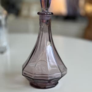 May include: A vintage, amethyst-colored glass perfume bottle with a faceted, geometric design. The bottle has a matching stopper and a flared base. The bottle is approximately 6 inches tall.