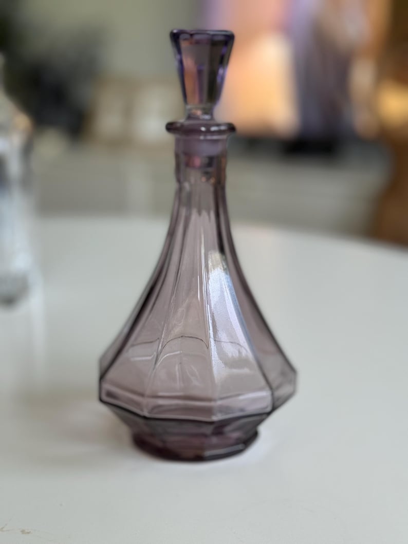 May include: A vintage, amethyst-colored glass perfume bottle with a faceted, geometric design. The bottle has a matching stopper and a flared base. The bottle is approximately 6 inches tall.
