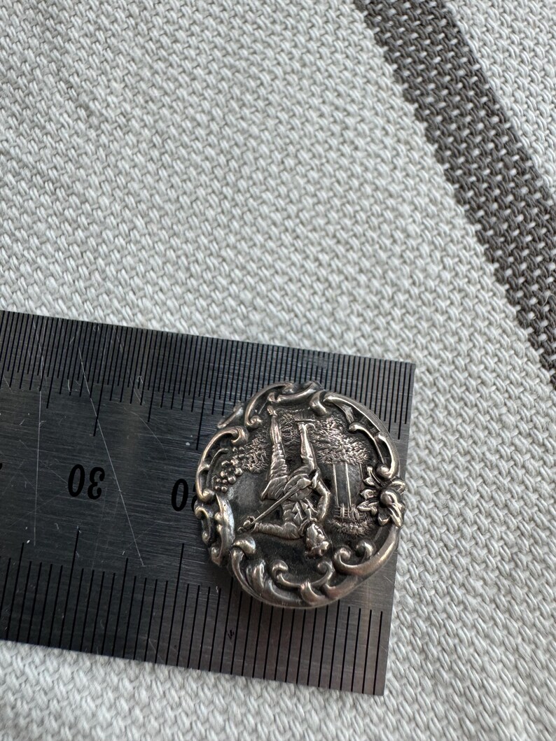 May include: Silver-toned button with an intricate design featuring a golfer swinging a club. The button is oval-shaped and has a detailed floral border.