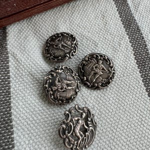 May include: Four silver-toned buttons with ornate designs featuring figures in various poses. The buttons have a raised, textured surface and intricate details.